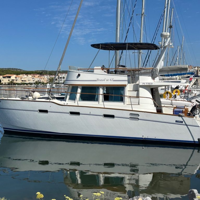 Bateau à moteur blanc amarré dans un port, avec un ciel bleu et des voiliers en arrière-plan.